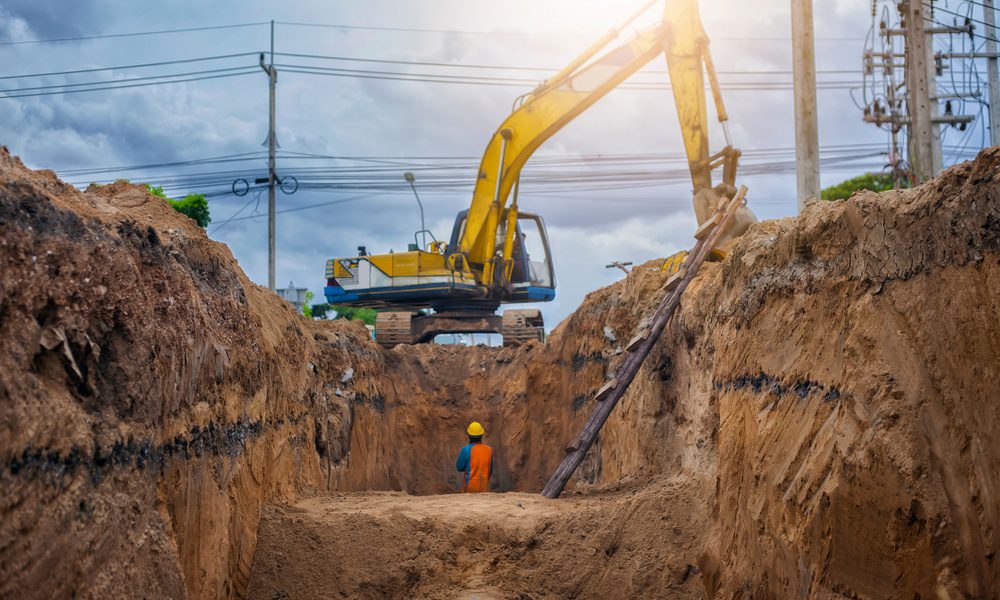 A yellow excavator works above a deep trench while a worker stands near a buried pipe and utility lines.