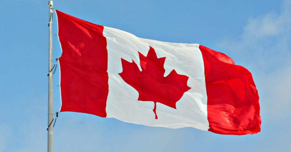 Canadian flag with a red maple leaf waving from a silver flagpole against a clear blue sky with light clouds.