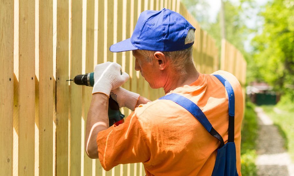 Fence installer in an orange shirt and blue cap uses a power drill to secure vertical boards on a wooden fence.