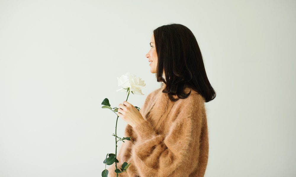 A woman in a beige sweater holding a white rose near her chest while facing left against a pale green background.