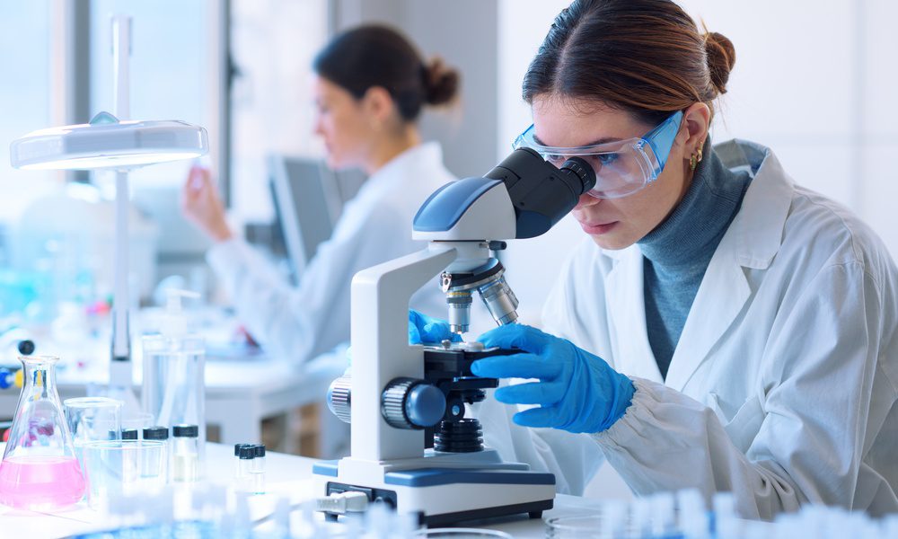 A laboratory scientist wearing safety glasses and gloves while examining a sample through a microscope.