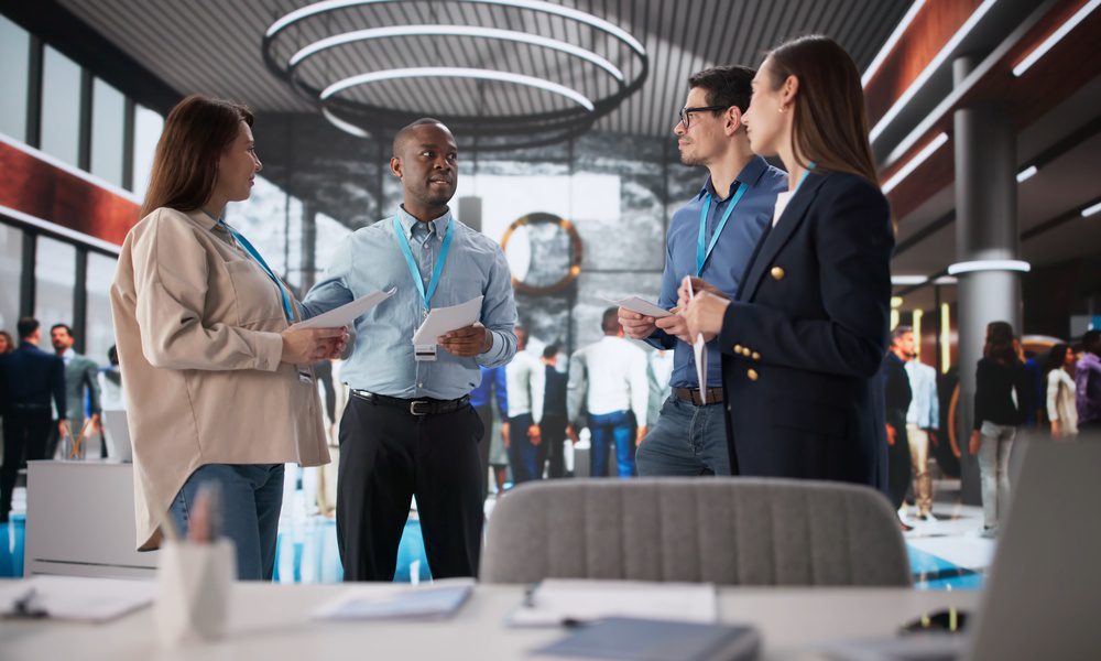 Four people wearing lanyards holding papers while talking in a busy conference hall with round ceiling lights.
