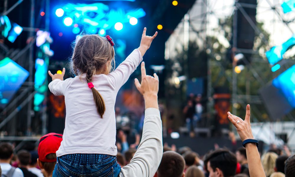 A close-up of a child sitting on someone's shoulders, raising both arms in front of a lit outdoor stage.