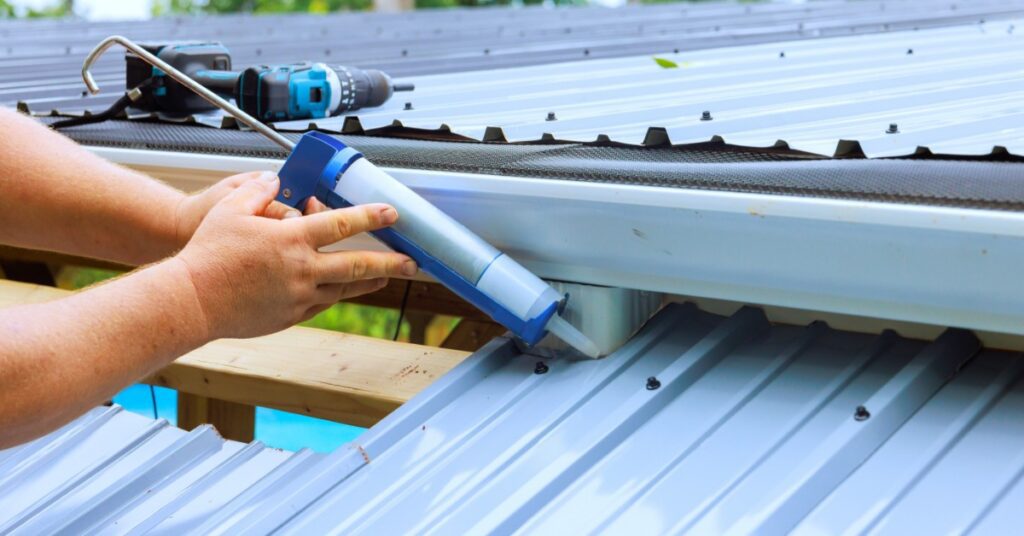 A close-up of the hands of a man using a glue gun on a beam poking through a metal roof next to large metal sheets.
