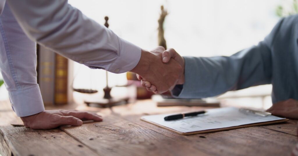 Close-up of two people shaking hands over a desk with paperwork, a pen, books, and scales of justice behind them.