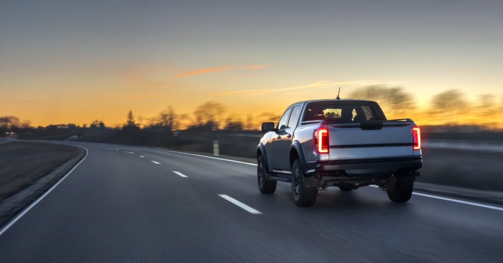 A gray pickup truck with brake lights on, driving on a concrete road with multiple trees on the side during a sunset.