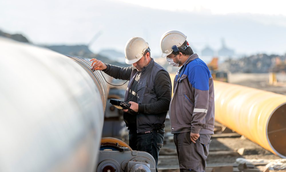 Two workers wearing white helmets, standing next to a large white pipe, while using an ultrasonic testing device.