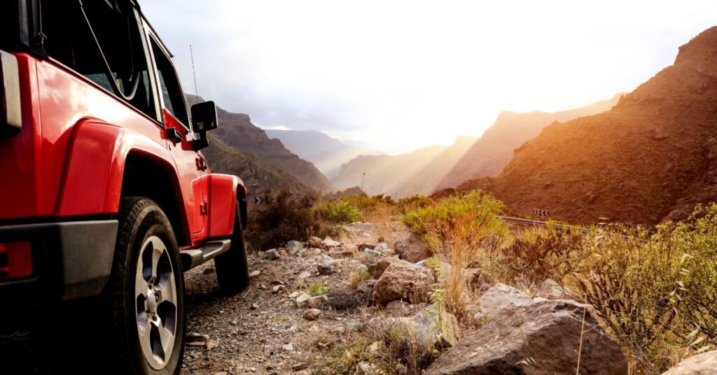 A close-up of the side of a red Jeep crossing through a dirt trail with multiple rocks in between dirt mountains.