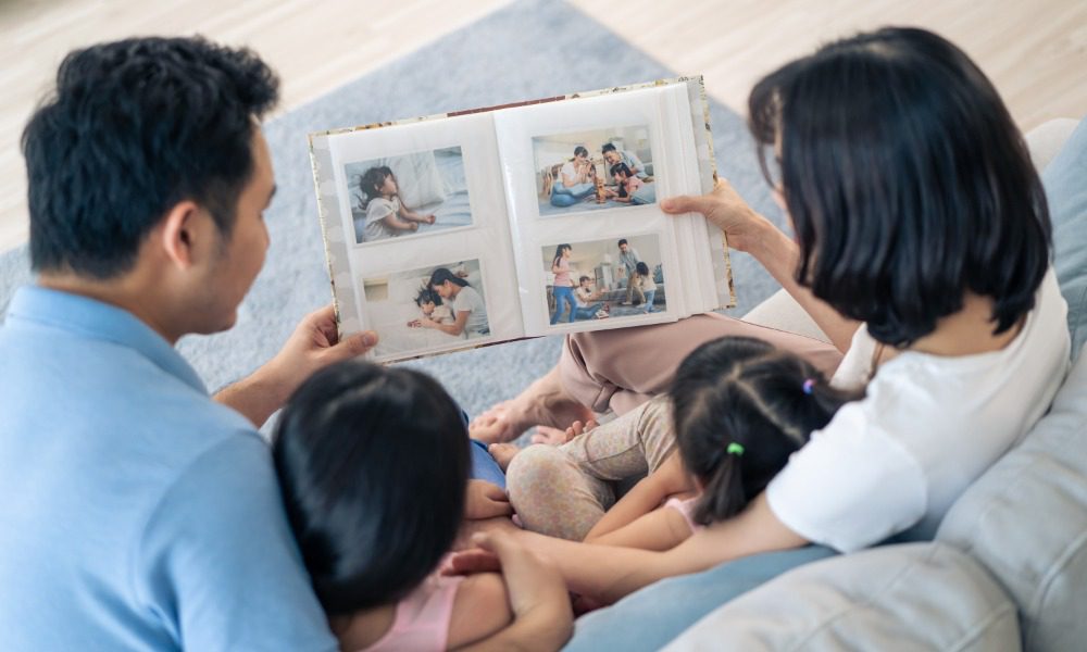 A family of four sitting close together on a gray sofa while the adults hold open an album filled with family photos.