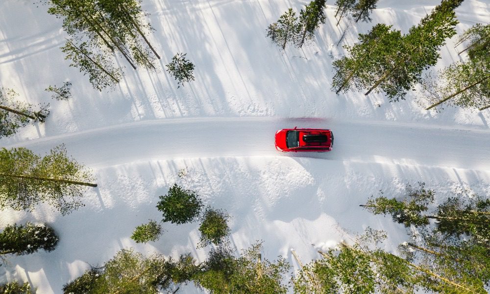 Bright red car driving past thin trees on a sunny day. Soft snow covers the road and surrounding terrain.