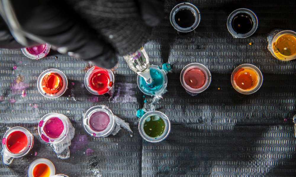 A person wearing black gloves dipping a tattoo pen into blue ink that is on a desk covered in different ink colors.